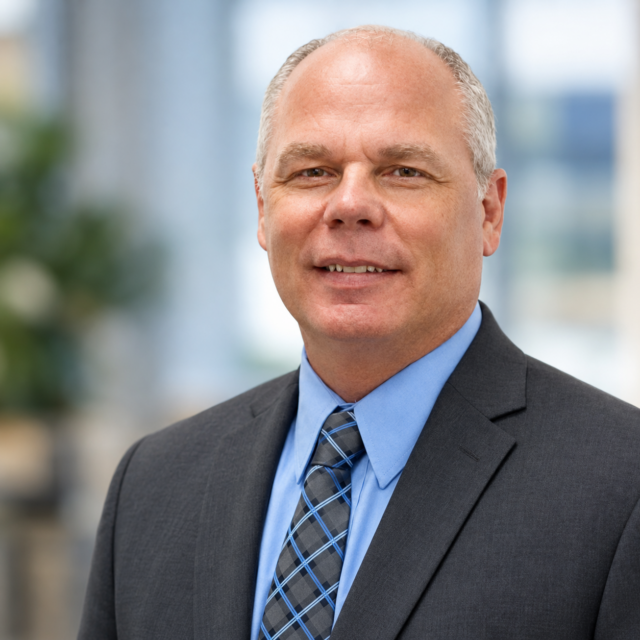Headshot of a smiling middle-aged man in a dark suit, light blue shirt, and blue-gray plaid tie, against a blurred background.