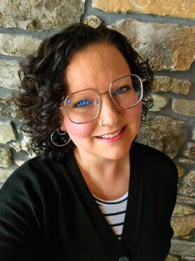 Smiling woman with curly dark hair and large round glasses, standing against a textured stone wall.